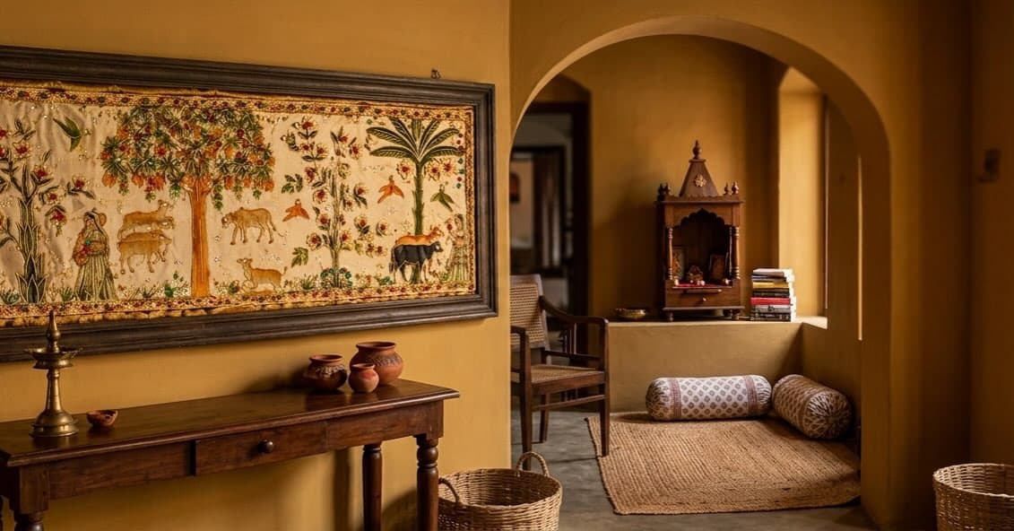 A wide framed textile panel hung on an ochre plaster wall, embroidered with flowering trees, birds, grazing cows and two standing human figures on a pale ground, with a floral border. Below it stands a dark wooden console table holding a brass lamp and small clay pots. To the right, an arched opening leads to an alcove with a wooden shrine, a stack of books, two bolsters and a woven mat on the floor.